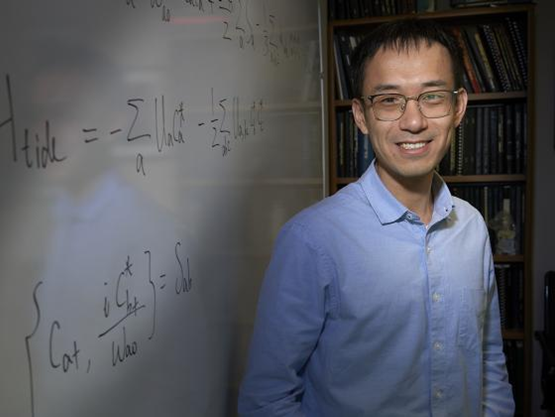 A person with short black hair and black-framed glasses wearing a blue button up shirt smiling to the camera