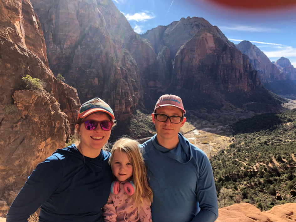 Two adults standing with a child between them with mountains and the sun in the background
